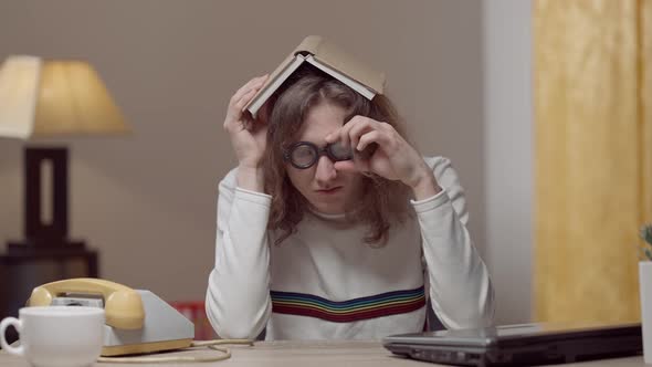 Tired Sad Retro Nerd in Eyeglasses Sitting at Table Indoors Holding Book on Head