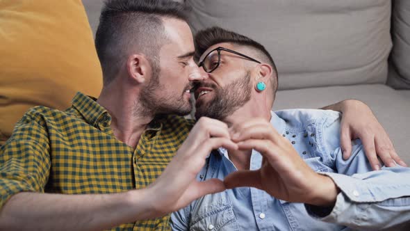 Young Gay Couple Kissing While Doing Heart Symbol with Hands at Home