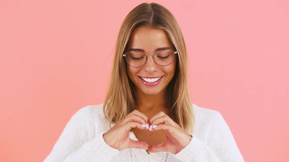 Young Female is Showing you a Heart Sign Made of Her Folded Fingers Smiling While Posing on Pink