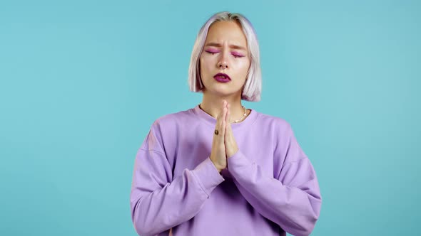 Cute Young Girl with Dyed Purple Hair Praying Over Blue Background. Woman Begging Someone Satisfy