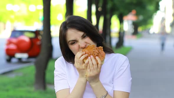 Brunette Woman Overeats on a Burger on the Street