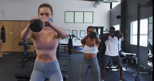 Two fit caucasian women and fit african american man wearing face masks exercising using kettlebell