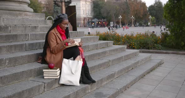 A Young Black Girl is Sitting on the Stairs on the Street and Reading a Book