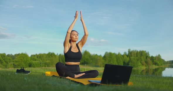 Young Focused Woman Does Yoga Exercises Using Laptop While Sitting on Mat on the Grass