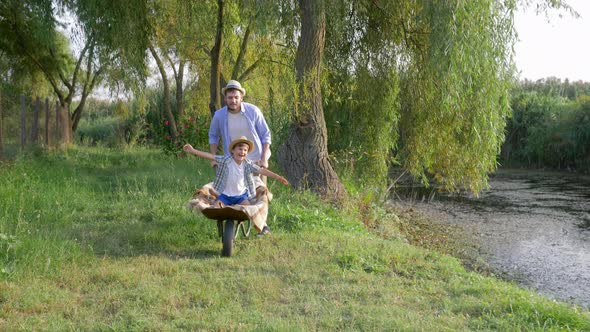 Cheerful Dad Fool Around with His Little Son in Wheelbarrow at Family Vacation in Rural