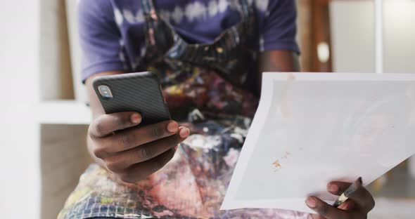 Midsection of african american male painter holding smartphone and photograph in artist studio