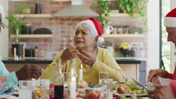 Group of happy diverse senior friends in santa hats talking at christmas dinner table at home