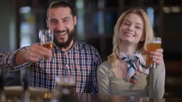 Front View Positive Man and Woman Toasting Looking at Camera Drinking Beer Sitting at Bar Counter