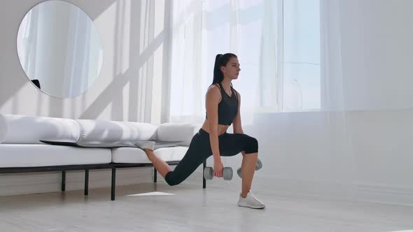 Brunette in Black Sportswear in a White Apartment Makes a Split Squat with Dumbbells in Her Hands