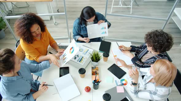 Group of Women Sharing Business Ideas Exchanging Papers with Charts Sitting at Desk Together