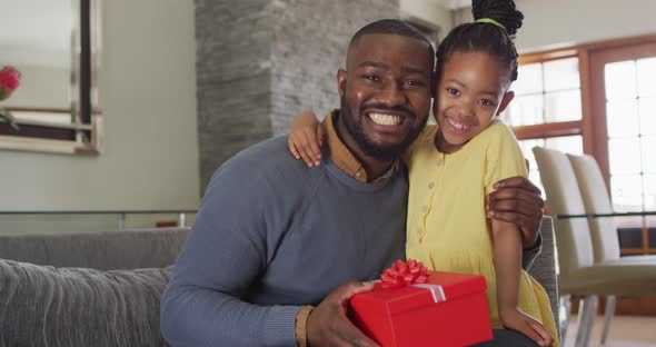 Happy african american daughter and father holding present sitting on sofa and looking at camera