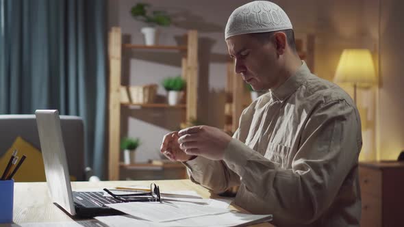 An Agitated Muslim in Skullcap Sits in the Living Room at a Laptop and Counts Finances Money Problem