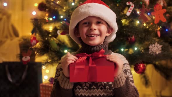 Portrait of Cheerful Smiling Boy with Christmas Present Box Wearing Santa Hat Sitting Next to