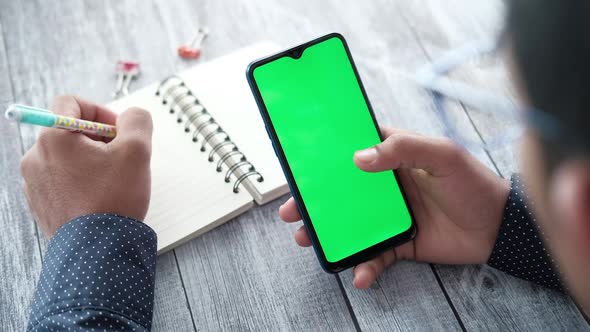 Rear View of Young Man Using Smart Phone and Writing on Notepad