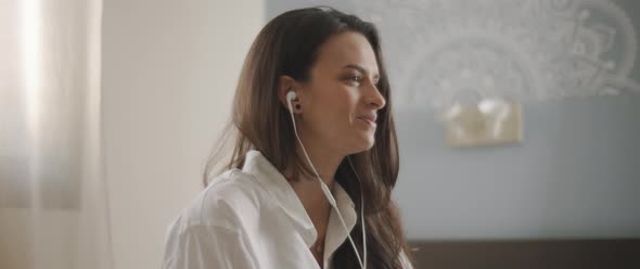 Close up of a young woman talking to someone through earphones while sitting on bed. 