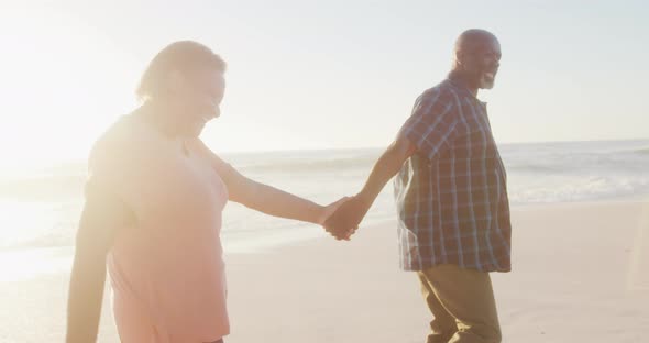 Smiling senior african american couple holding hands and walking on sunny beach