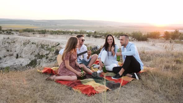 Four Friends Cheering with Beer and Smiling While Sitting on the Blanket at a Summer Picnic