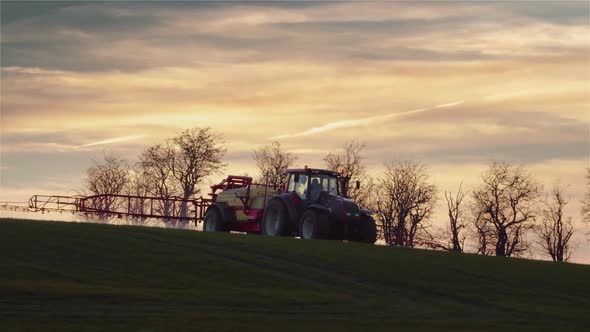 A farmer with a tractor spraying chemicals to a field during sunset. Panning shot.