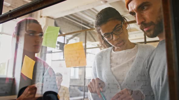 Three Young Business People Discussing and Planning Strategy in Front of Glass Board and Stickers