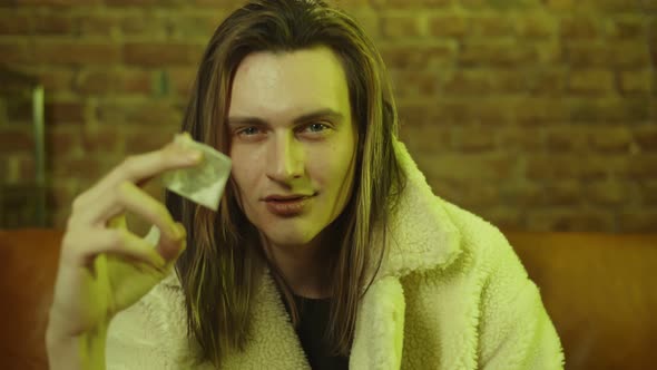 Young Man Drug Dealer in Coat Sitting on Couch and Holding Heroine in Bag Looking at Camera