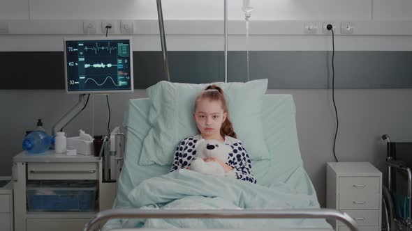 Portrait of Hospitalized Sick Girl Child Patient Holding Teddy Bear Resting in Bed During Medical