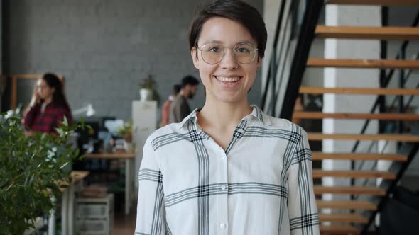 Portrait of Happy Young Lady Standing in Office Smiling While People Working in Background