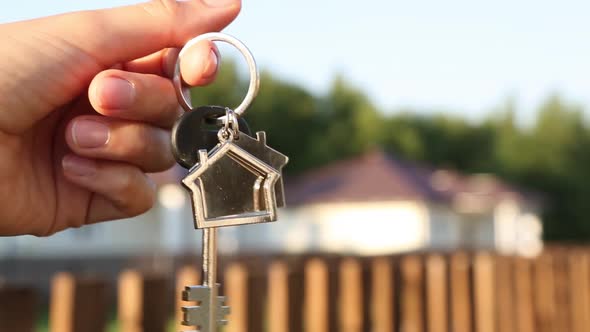 Key ring with keychain house in hand. Background of fence and cottage