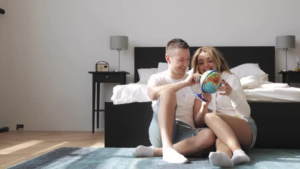 Young Couple Sitting Beside the Bed Are Looking at Globe Choosing a Place To Travel on Honeymoon