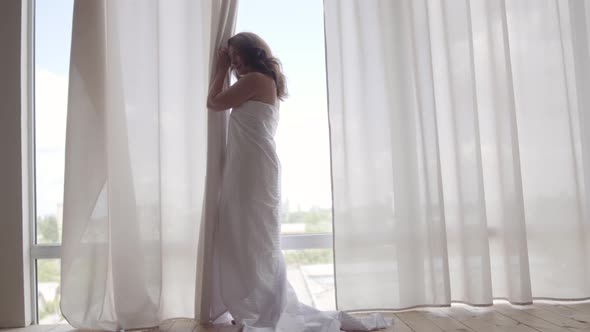 Attractive Woman Covered in the Bedsheet Standing Near the Floor-to-ceiling Window and Looking Away