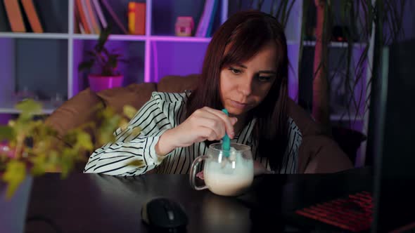 Young Woman Stirs Coffee with Spoon Sitting in Armchair Before Computer in Office