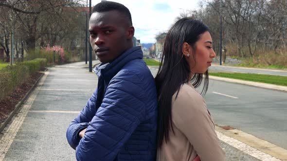 A Black Man and a Asian Woman Stand in a Street with Their Backs Toward Each Other and Frown