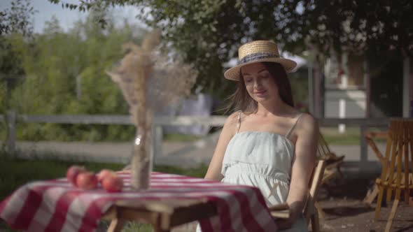 Young Rural Girl in a Straw Hat and White Dress Sitting at the Small Table with Vase with Decorative