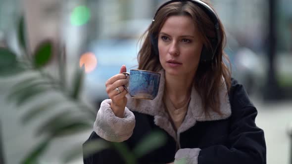 Thoughtful Woman Is Enjoying Coffee and Music By Headphone, Sitting in Cafe on Street Terrace