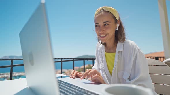 Young Elegant Woman with Wireless Earphones Looking at Laptop with Smile While Sitting at Balcony