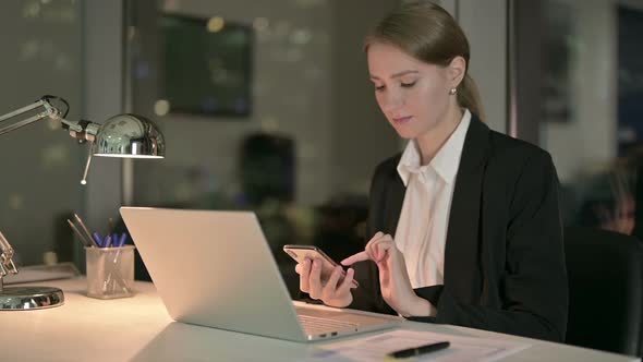 Young Businesswoman Scrolling on Cellphone in Office at Night