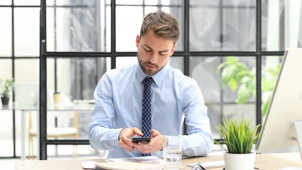 Happy businessman holding smartphone texting message at office, receiving sms message reading good n