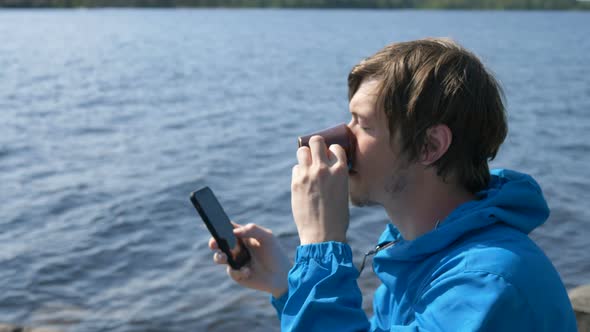 Hiker Drinks Coffee From Small Cup Using Phone Near River