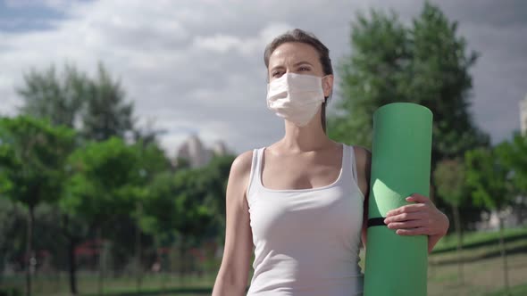 Confident Caucasian Sportswoman Walking with Exercise Mat Outdoors. Taking Off Covid-19 Face Mask