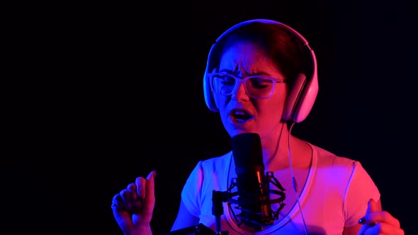 Caucasian Woman in Glasses and Headphones Sings Into a Microphone in Neon Light on a Black