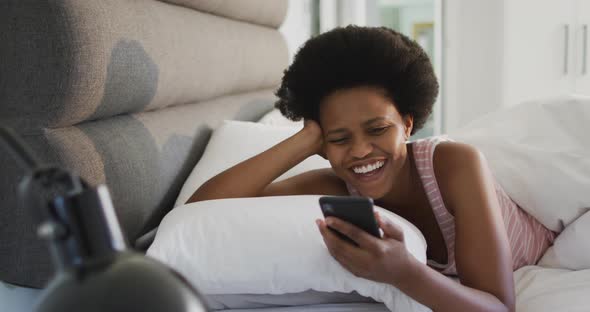 Happy african american woman laying in bed, using smartphone in bedroom