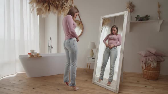 Displeased Lovely Adult Woman Trying to Button Tight Denim in Front of Mirror After Gaining Weight
