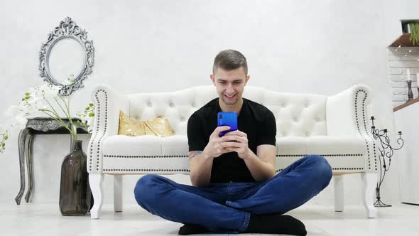 Young Man Sitting on the Floor in Modern Luxury House and Using Smartphone, Chatting with Friends