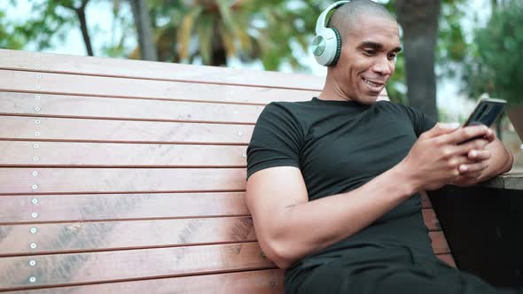 Cheerful bald African man typing on phone in headphones while sitting on the bench