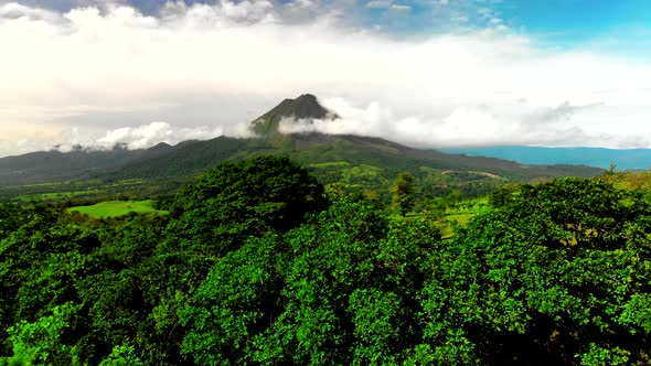 Drone moves up revealing beautiful and green volcano Arenal behind the trees.
