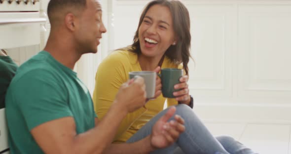 Happy biracial couple sitting on kitchen's floor, drinking coffee and talking