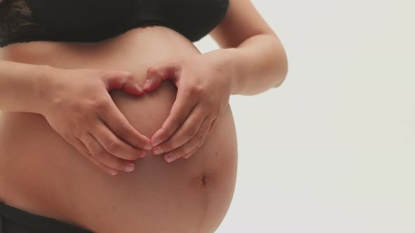 A Pregnant Girl Holds Her Hands on Her Bare Stomach in the Shape of a Heart on a White Background