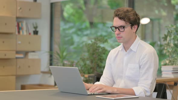 Young Man Looking at Camera While Using Laptop in Modern Office