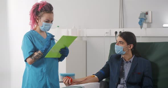 Nurse in Uniform and Safety Mask Using Clipboard and Taking Blood Sample From Patient Lying on Couch