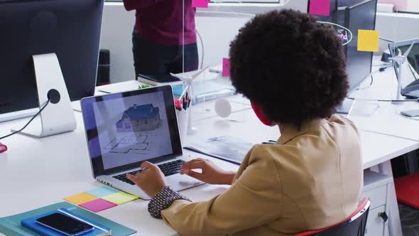 Mixed race businesswoman wearing face mask sitting using a laptop in modern office