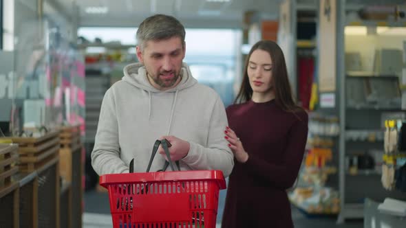 Slim Young Woman Approaching to Man Looking Through Supplies in Shopping Basket